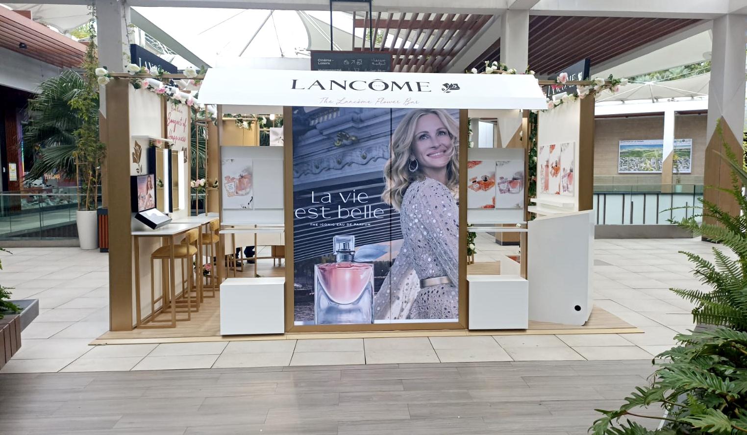 Flower bar Lancôme « La vie est belle » : structure en bois clair, auvent blanc et guirlandes de fleurs.